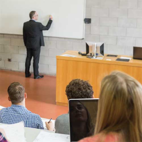 Picture of a lecture hall with instructor at whiteboard and students taking notes.