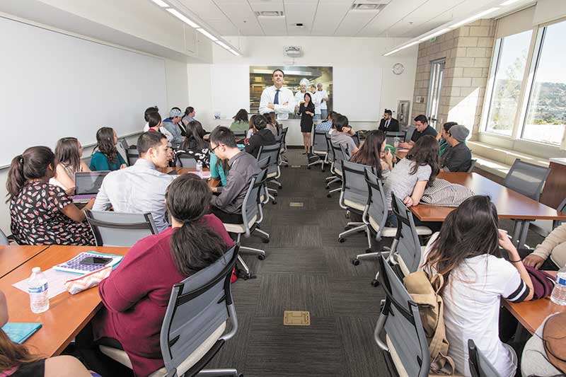 Shot from back of classroom full of students. Teacher at front in front of IDEA surface with projection on it.