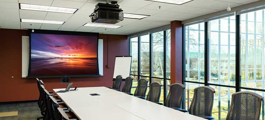 Conference room with a large table and chairs, projector screen and projector mount, in-wall PTZ camera and ceiling microphones.